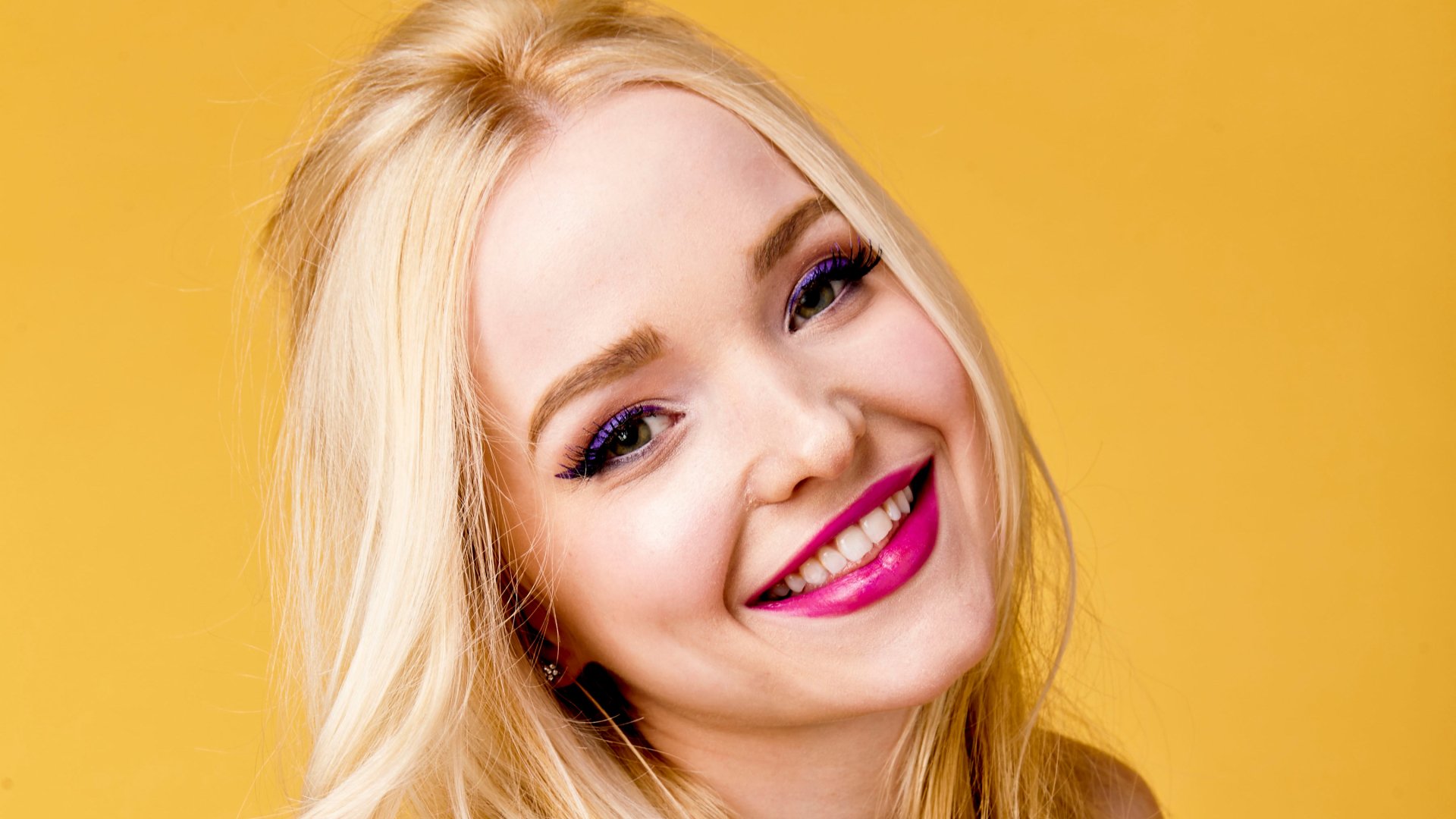 Close-up HD desktop wallpaper of blonde celebrity Dove Cameron smiling against a warm yellow background.