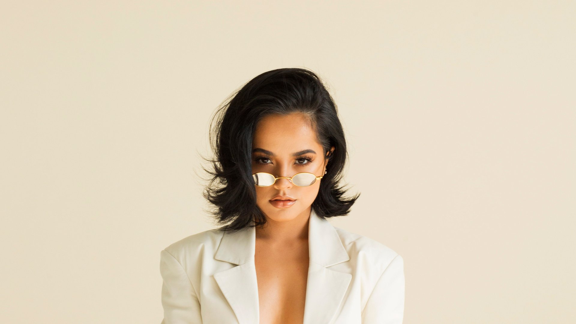 HD desktop wallpaper featuring American singer Becky G with black hair, brown eyes, and stylish sunglasses against a neutral background.