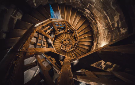 Top-down view of a man-made spiral wooden staircase winding inside stone walls; HD PC desktop wallpaper and background featuring rustic stairs, warm tones, and intricate geometric patterns.