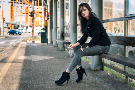 A woman with black hair and high heels sits on a bench at a bus stop, with a blurred urban background captured in HD with a shallow depth of field.