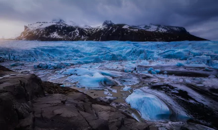 2K Quad HD PC desktop wallpaper: nature scene of a bright blue glacier flowing through a rocky foreground toward snow-capped mountains beneath a dramatic cloudy sky.