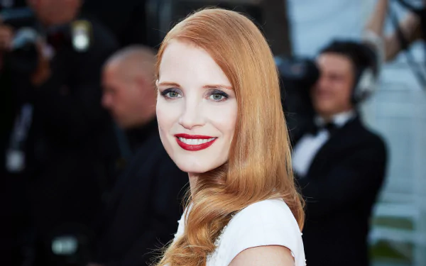 HD desktop wallpaper featuring American actress Jessica Chastain with red hair, blue eyes, red lipstick, and a bright smile at a formal event.