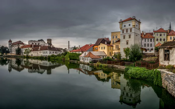 8K Ultra HD desktop wallpaper featuring a serene man-made lake reflecting the charming buildings of a historic town under a cloudy sky.
