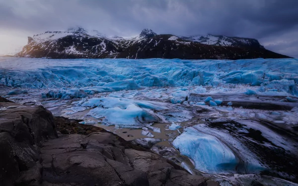 2K Quad HD PC desktop wallpaper: nature scene of a bright blue glacier flowing through a rocky foreground toward snow-capped mountains beneath a dramatic cloudy sky.