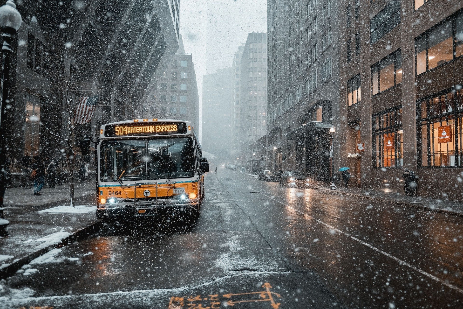HD desktop wallpaper showing a city street with a bus stopped amid heavy snowfall, wet roads, and tall buildings lining the street.