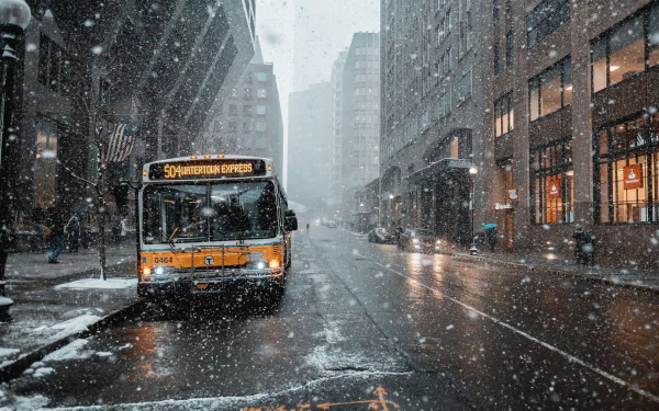 HD desktop wallpaper showing a city street with a bus stopped amid heavy snowfall, wet roads, and tall buildings lining the street.