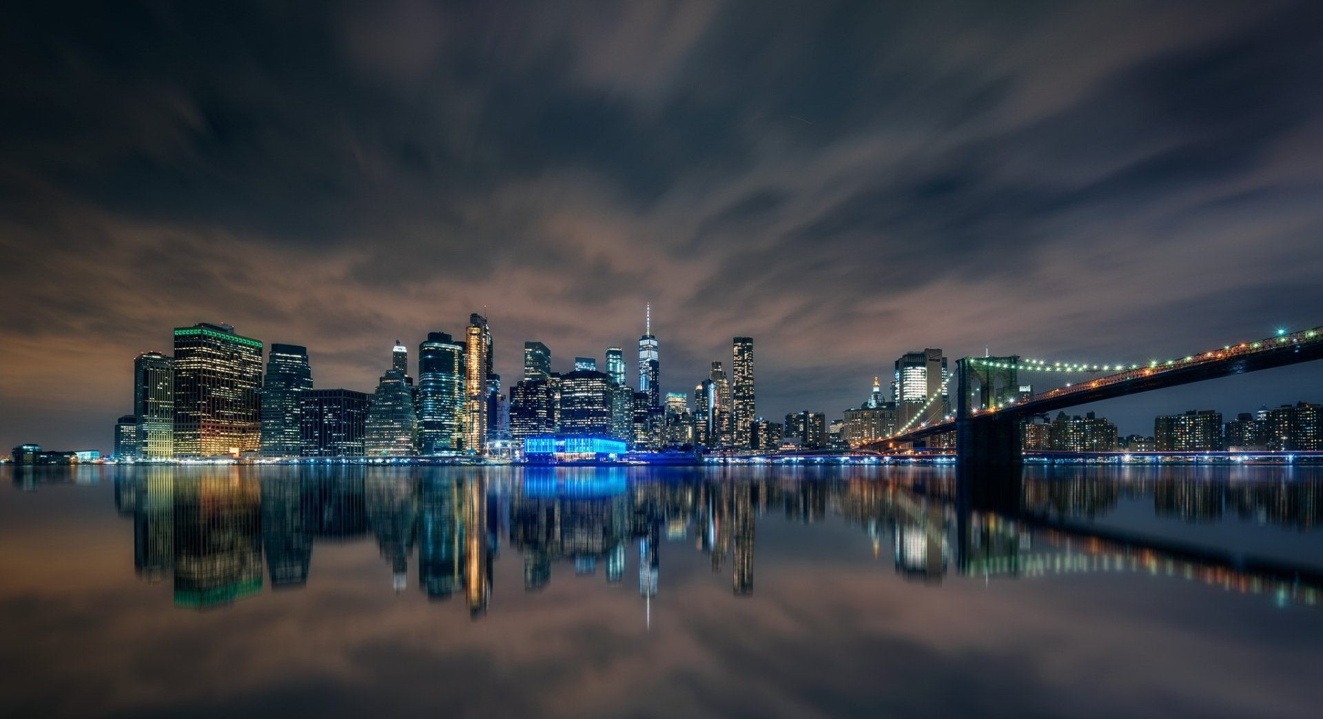 Night view of Manhattan's skyline with the Brooklyn Bridge and skyscrapers reflected in the water, showcasing New York City's vibrant urban landscape.