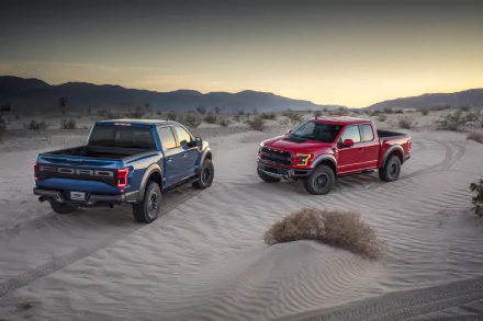 8K Ultra HD PC desktop wallpaper: two Ford F-150 Raptor trucks (blue and red) parked on windswept desert dunes at sunset.