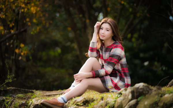 A smiling Asian brunette woman in a plaid shirt sits outdoors on a rock, with a blurred natural background captured in 4K Ultra HD depth of field.