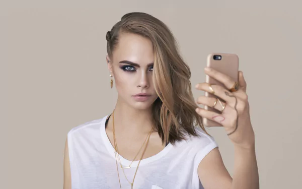 Close-up of blonde English model and actress Cara Delevingne with striking blue eyes holding a smartphone, captured in 4K Ultra HD quality for desktop wallpaper.
