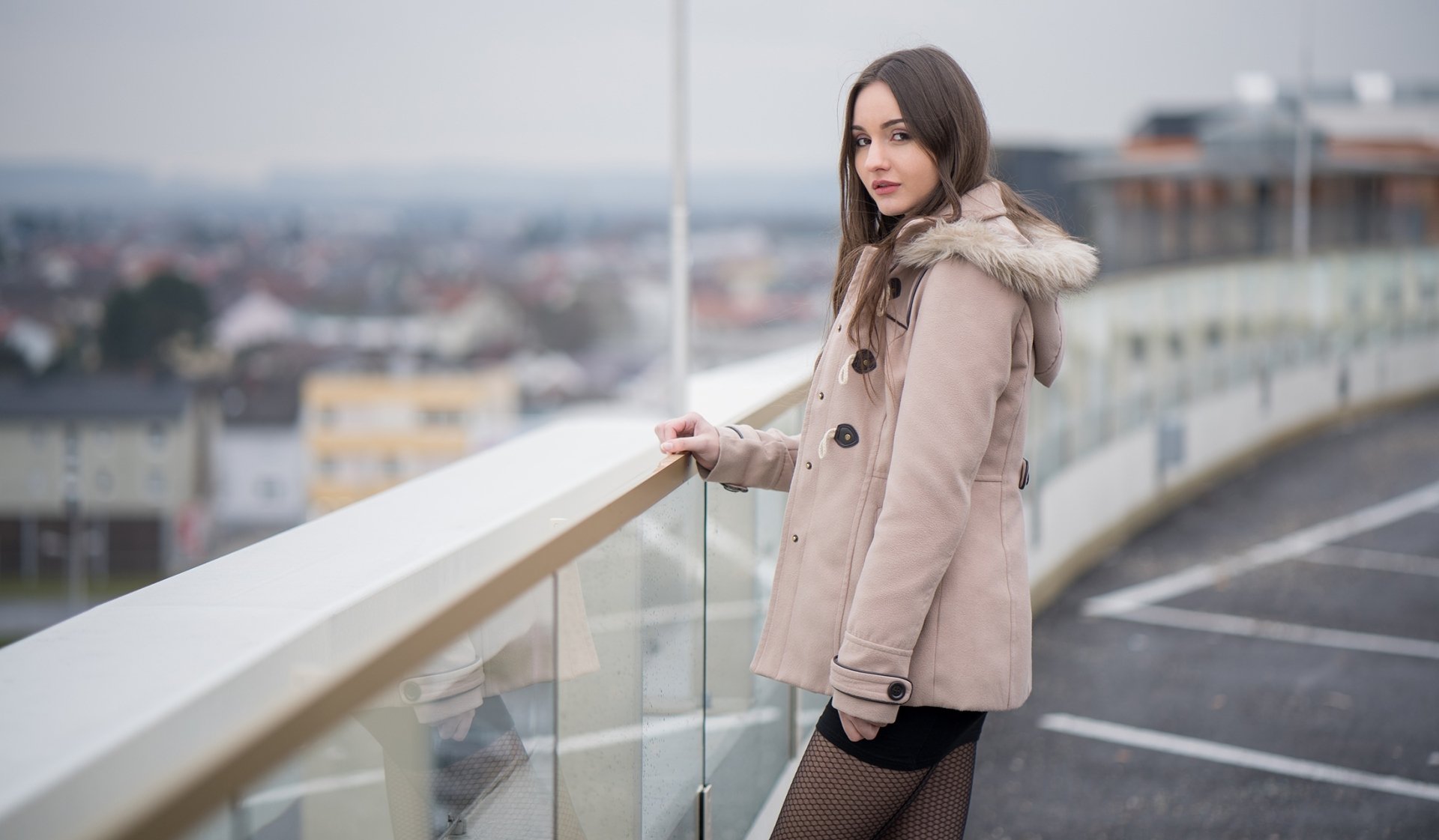 Brunette woman wearing a jacket stands on a rooftop with a cityscape blurred in the background, captured with a depth of field effect in this HD desktop wallpaper.
