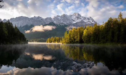 HD desktop wallpaper capturing a serene Bavarian mountain landscape with a calm lake reflecting snow-capped peaks and lush autumn trees under a partly cloudy sky.