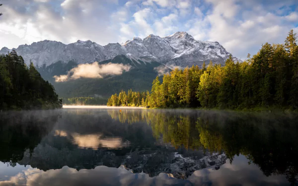 HD desktop wallpaper capturing a serene Bavarian mountain landscape with a calm lake reflecting snow-capped peaks and lush autumn trees under a partly cloudy sky.