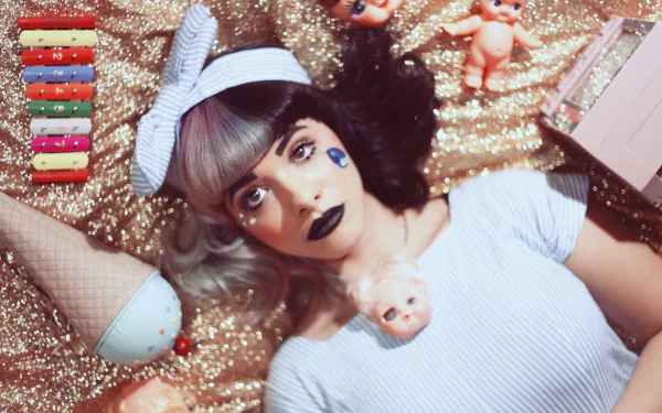 HD desktop wallpaper of American singer Melanie Martinez with bold lipstick, lying on a glittery surface surrounded by quirky dolls and colorful makeup items.