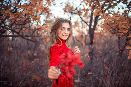 A blonde woman with blue eyes smiles warmly, extending red leaves toward the camera, set against a blurred autumn forest backdrop in 4K Ultra HD quality.