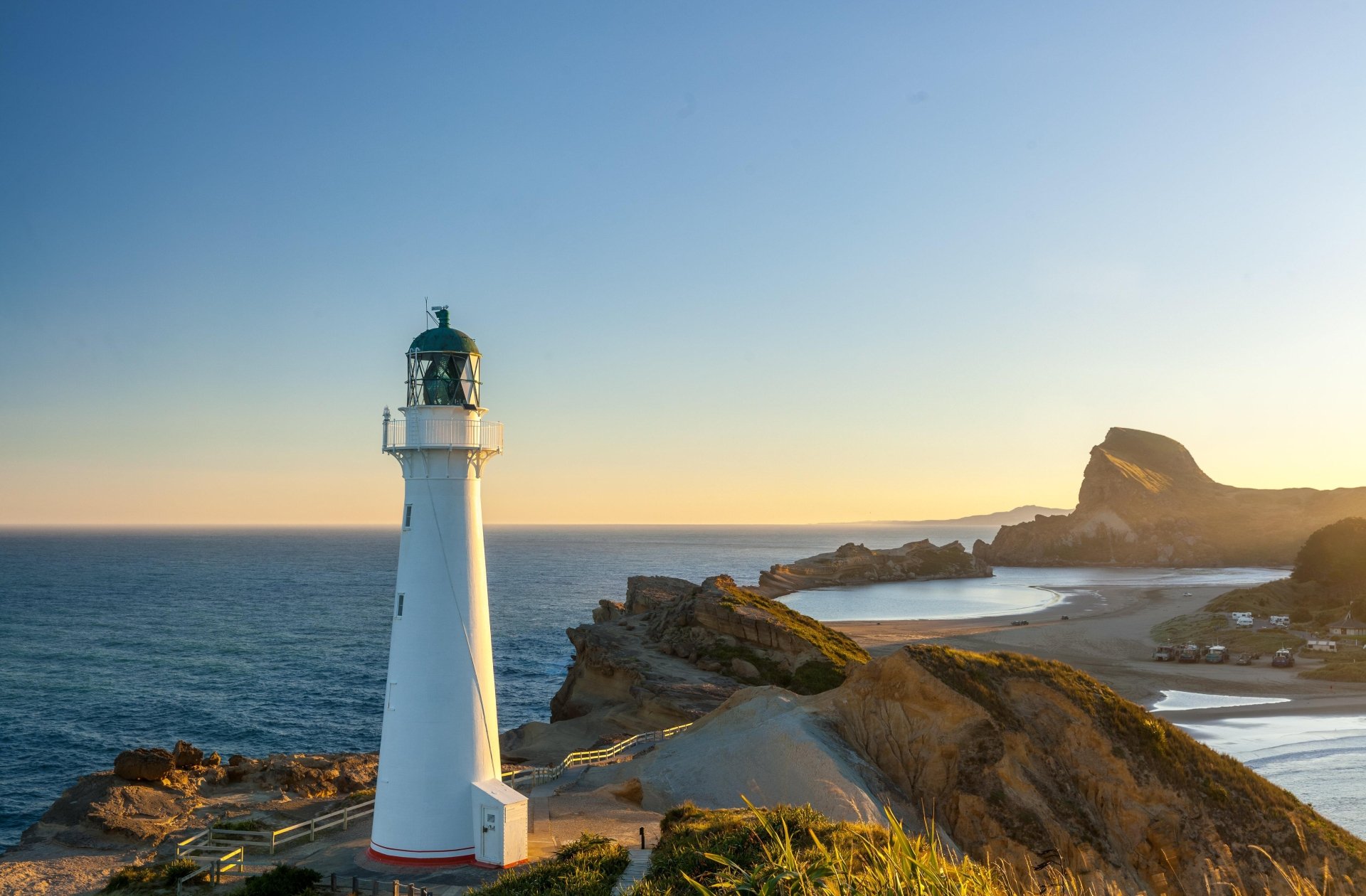 4K Ultra HD image of a man-made lighthouse on the New Zealand coast at horizon during sunset, featuring rugged cliffs and calm sea waters.