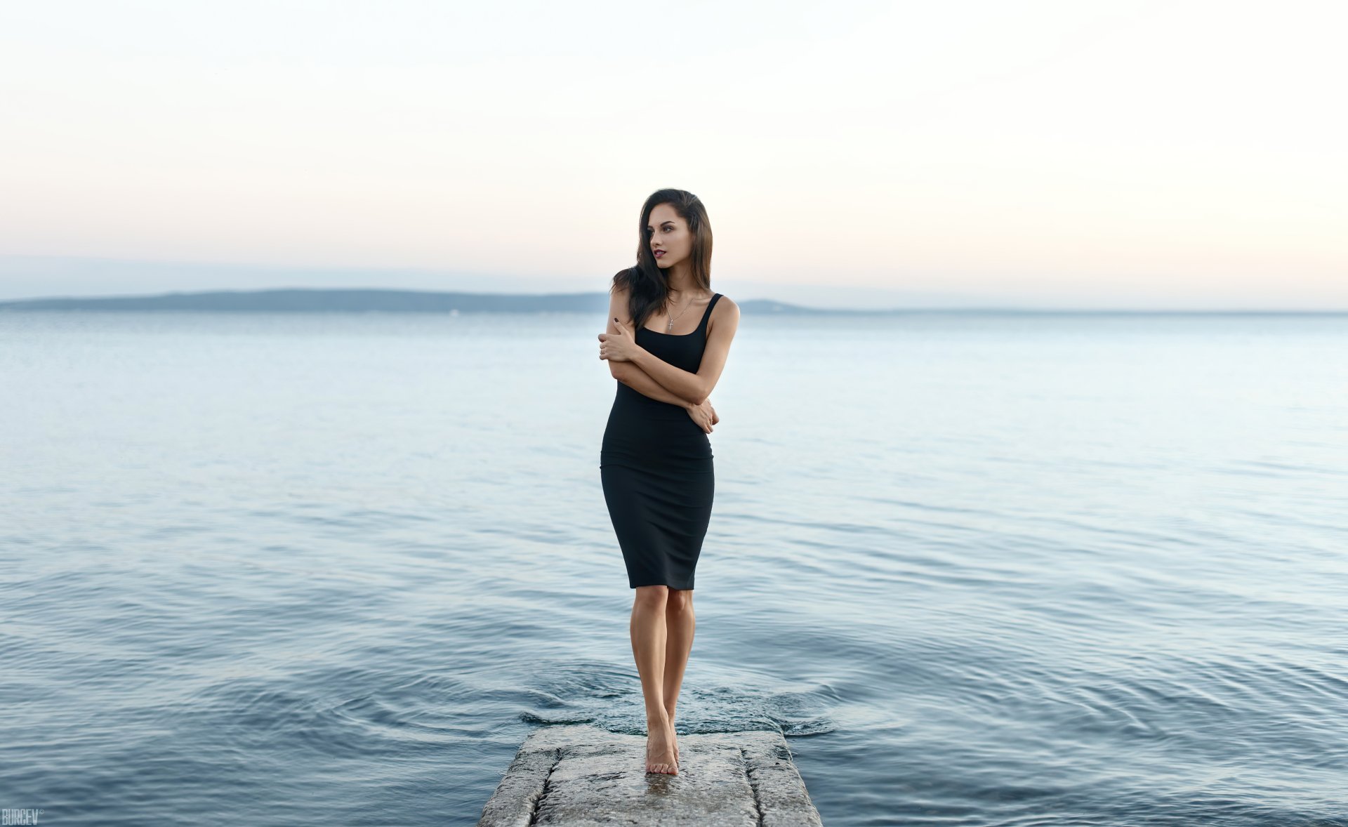 Brunette woman in a black dress stands poised on a pier with a blurred ocean backdrop, captured with striking depth of field in this 4K Ultra HD desktop wallpaper.