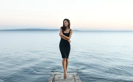 Brunette woman in a black dress stands poised on a pier with a blurred ocean backdrop, captured with striking depth of field in this 4K Ultra HD desktop wallpaper.