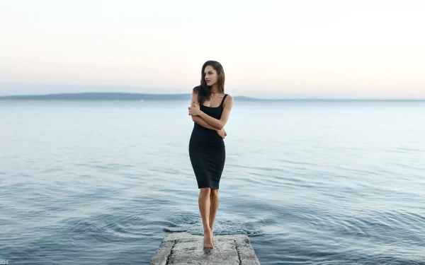 Brunette woman in a black dress stands poised on a pier with a blurred ocean backdrop, captured with striking depth of field in this 4K Ultra HD desktop wallpaper.