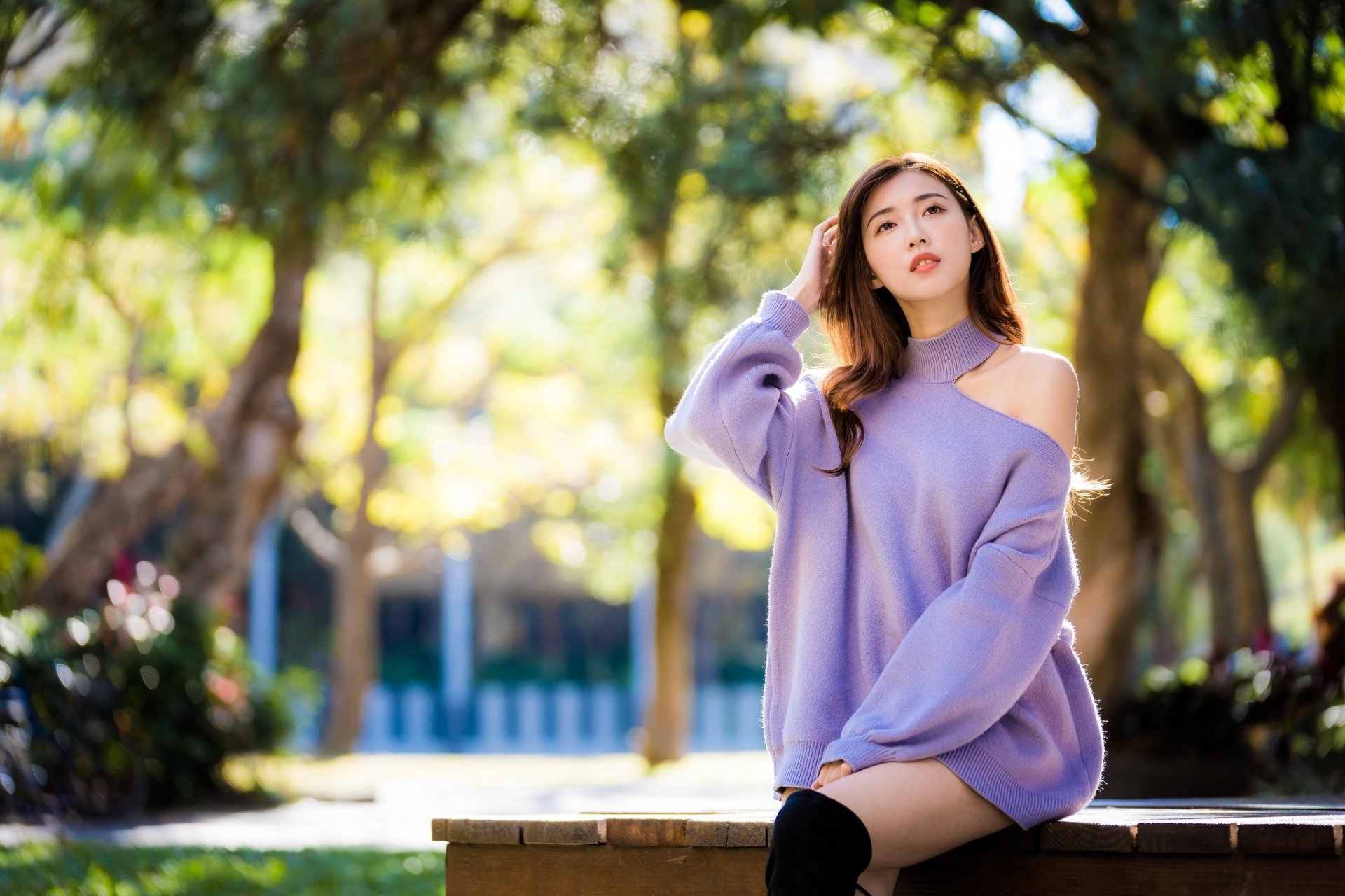 Asian woman with brunette hair wearing a lavender sweater, sitting outdoors with blurred trees in the background, captured with a depth of field effect in this HD wallpaper.