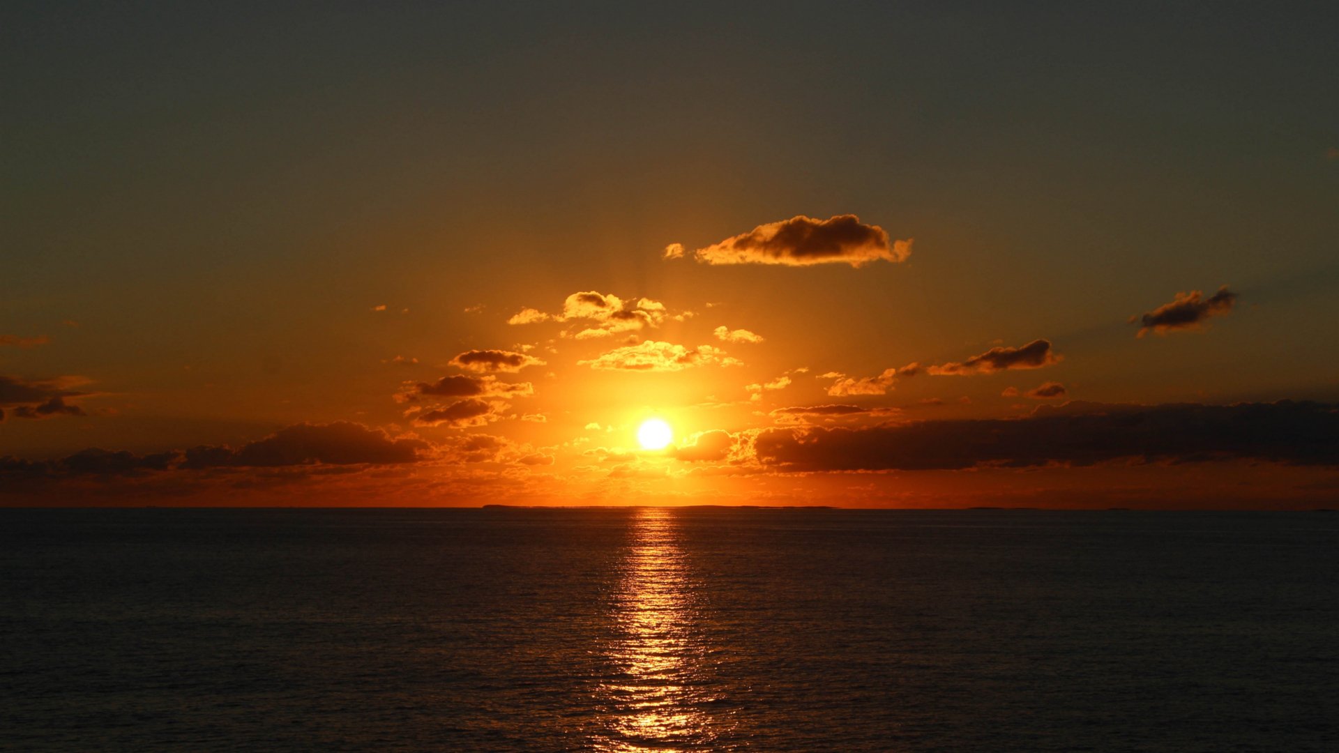 HD PC desktop wallpaper showing a serene sunset over the ocean with vibrant orange and yellow hues illuminating the sky and reflecting on the water.