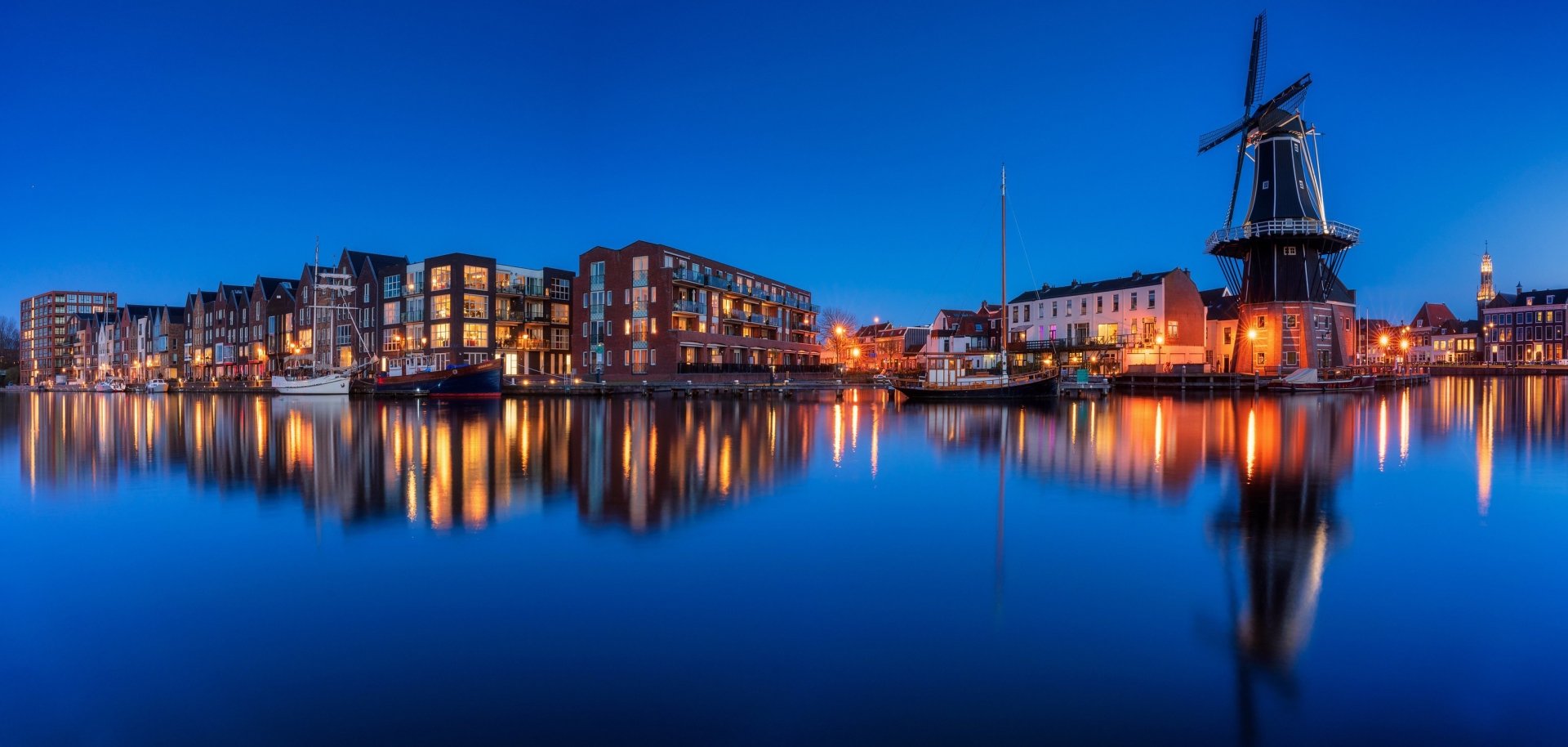 HD desktop wallpaper of Haarlem’s waterfront at dusk, featuring illuminated modern buildings and a traditional windmill reflecting on calm water under a deep blue sky.
