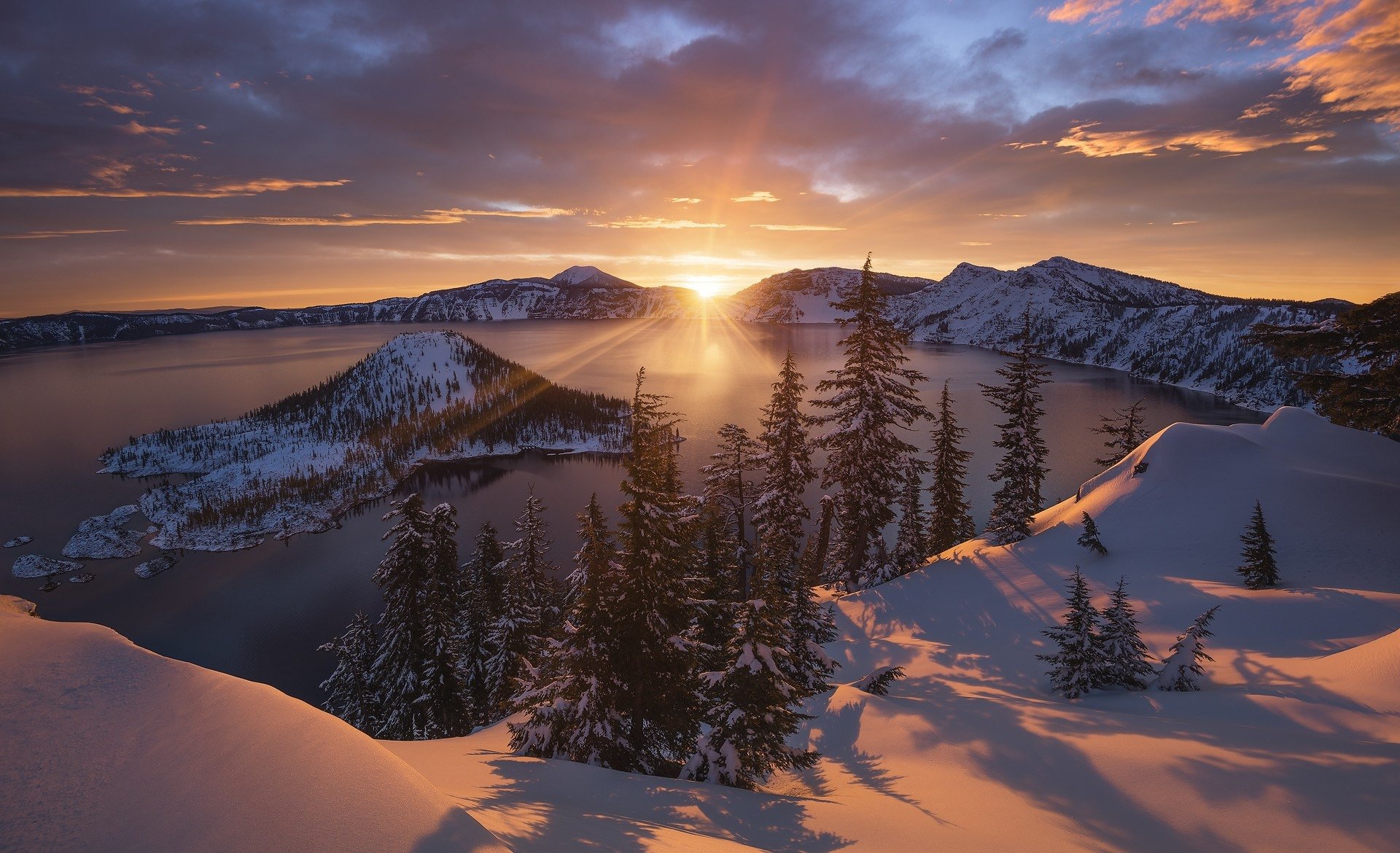 Sunbeam breaks through clouds over snowy Crater Lake in winter, with a vibrant sky illuminating the serene natural landscape.