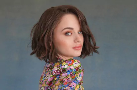 HD desktop wallpaper featuring American actress Joey King with blue eyes and short brunette hair, wearing a colorful floral top against a muted gray background.