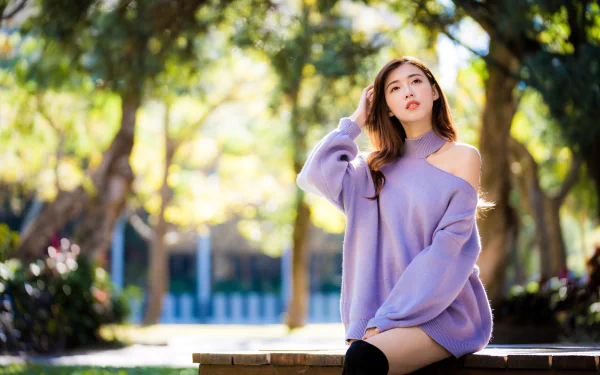 Asian woman with brunette hair wearing a lavender sweater, sitting outdoors with blurred trees in the background, captured with a depth of field effect in this HD wallpaper.
