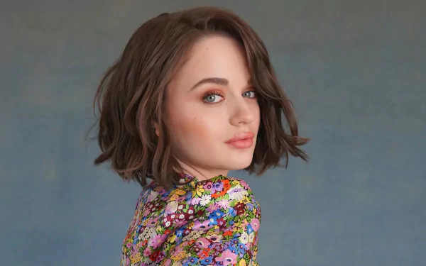 HD desktop wallpaper featuring American actress Joey King with blue eyes and short brunette hair, wearing a colorful floral top against a muted gray background.