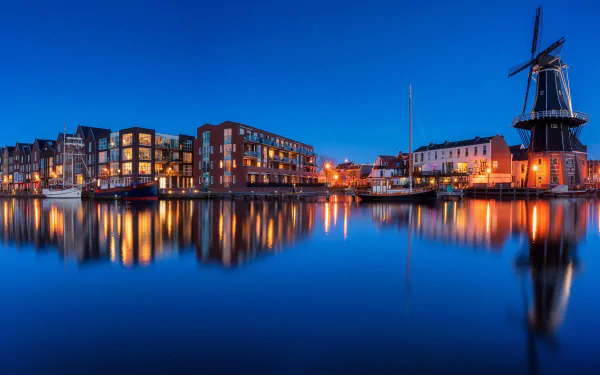 HD desktop wallpaper of Haarlem’s waterfront at dusk, featuring illuminated modern buildings and a traditional windmill reflecting on calm water under a deep blue sky.