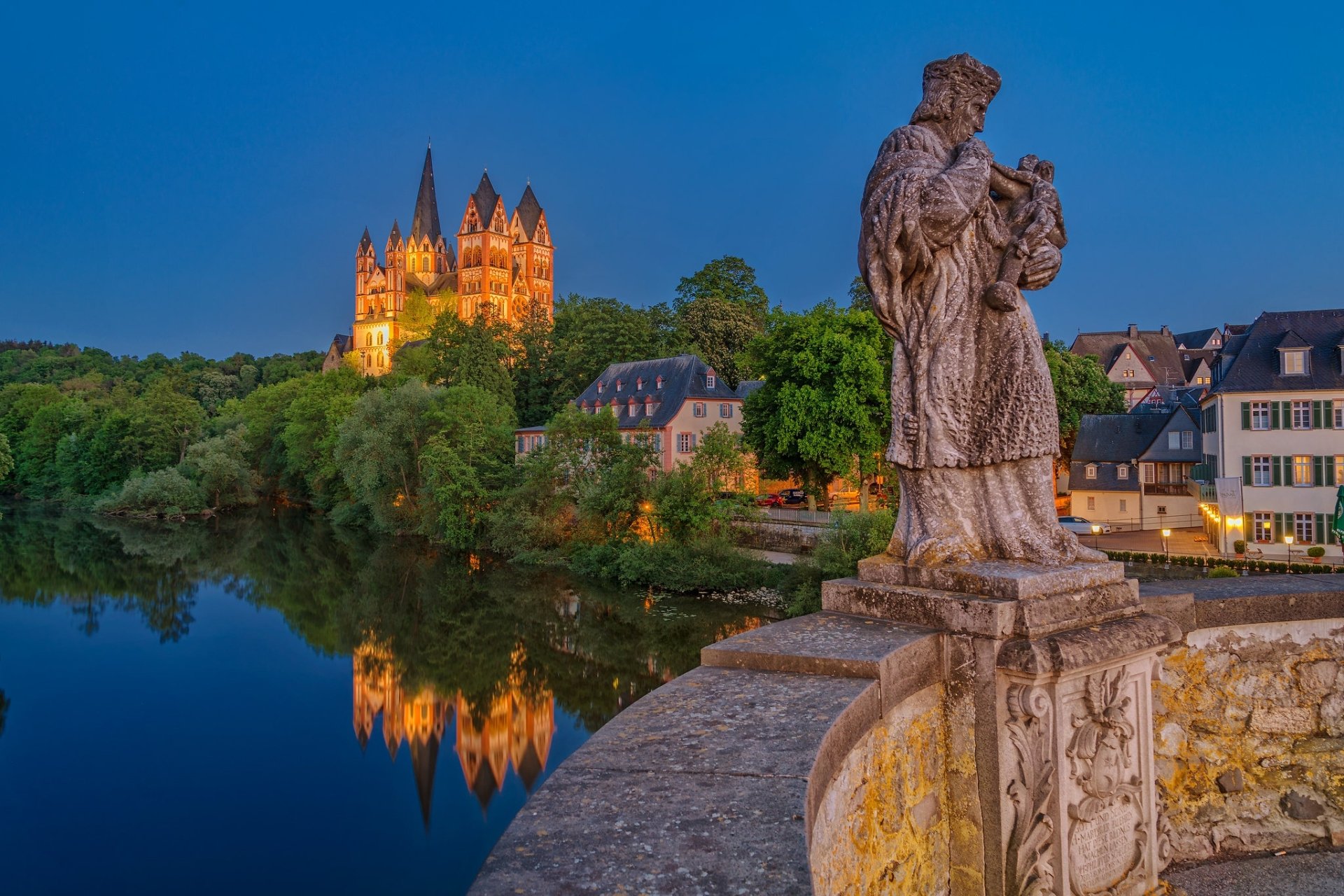 HD desktop wallpaper showing a religious statue overlooking a river with houses and a cathedral reflecting in the water during twilight.