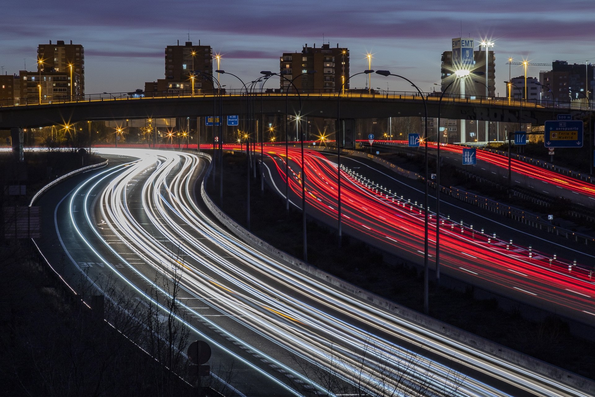 Time-lapse 4K Ultra HD photo of a busy Madrid road with light trails, city buildings, and a bridge under dusk skies in Spain.