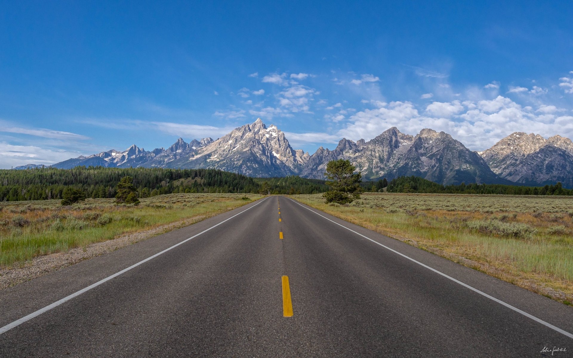 HD PC desktop wallpaper/background: a man-made paved road stretching through a grassy nature landscape toward jagged mountains beneath a blue sky.