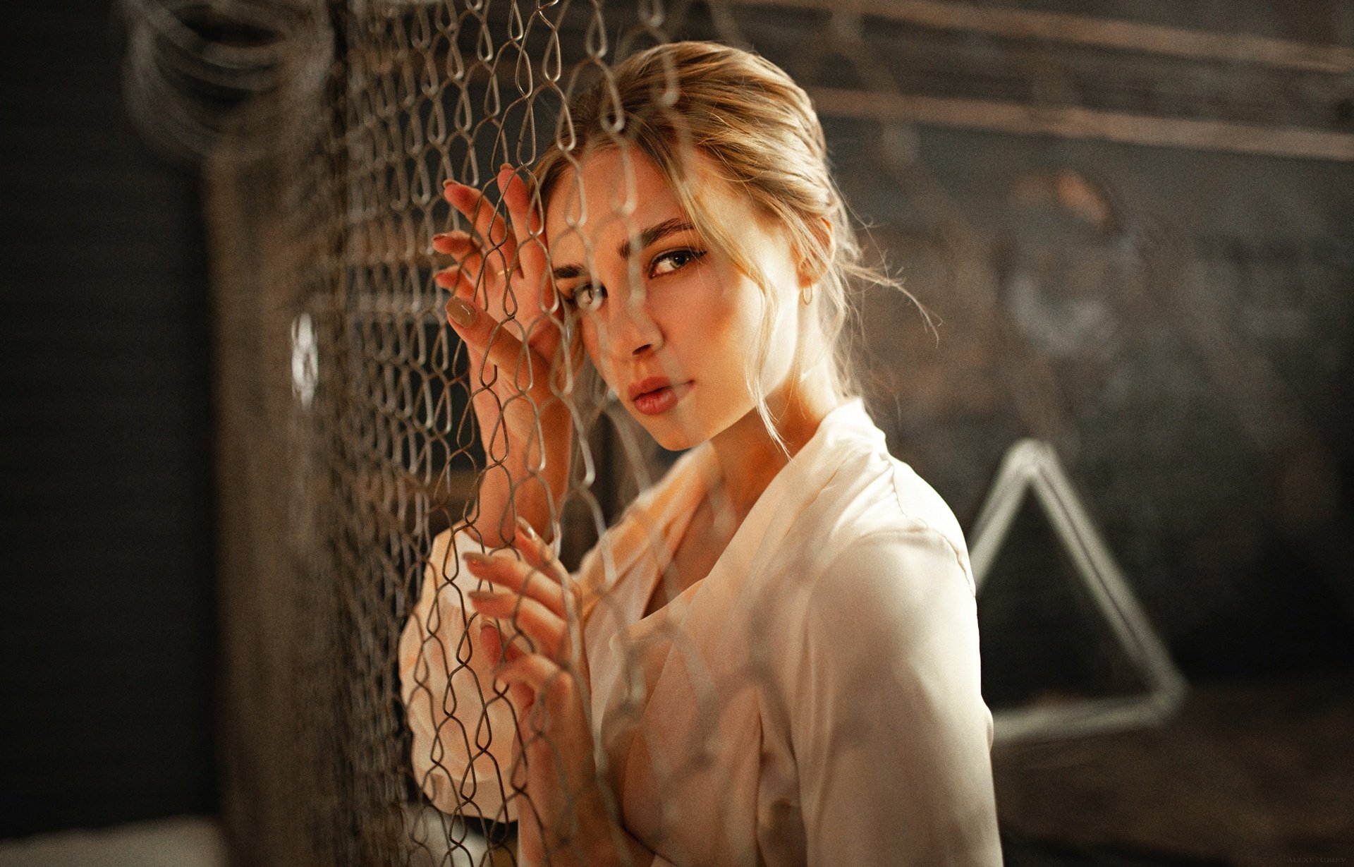 HD PC desktop wallpaper: portrait of a woman model, close-up of her face and hand on a chain-link fence, soft moody lighting with a blurred industrial background.