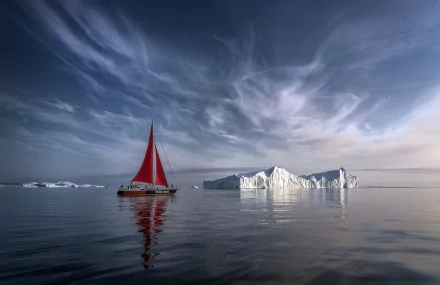 Sailboat with a red sail near a large iceberg in calm Arctic waters of Greenland under a dramatic sky, captured in HD for a desktop wallpaper background.