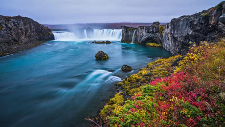 HD PC desktop wallpaper showcasing Iceland’s Goðafoss waterfall surrounded by rugged cliffs and vibrant autumn foliage under a soft, cloudy sky.