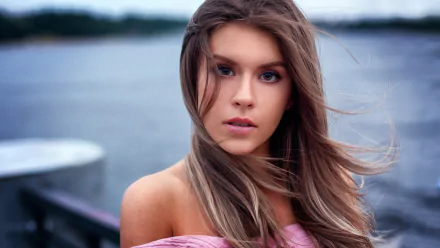 HD PC desktop wallpaper: portrait of a young woman model with long windblown hair, clear facial features and soft makeup, gazing at the camera against a blurred lakeside background.