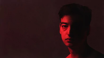 Moody HD desktop wallpaper: close-up portrait of a young male singer half-lit in red shadow against a dark, minimalist music-themed background.