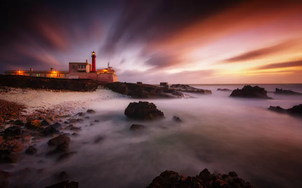 5K Ultra HD PC desktop wallpaper/background: Portuguese coastal lighthouse at sunset, dramatic clouds and misty waves over rocky shore — man-made beacon glowing against a vibrant sky.