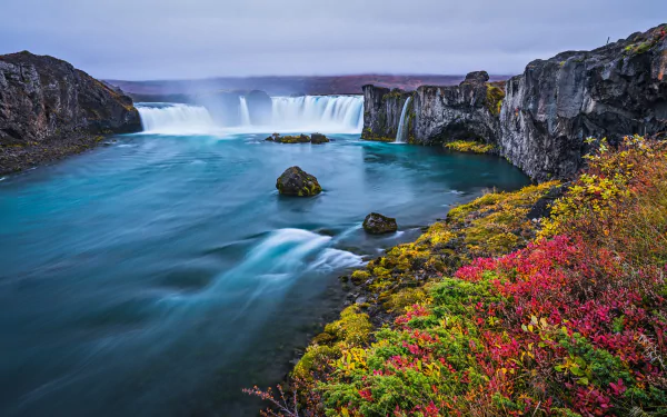 HD PC desktop wallpaper showcasing Iceland’s Goðafoss waterfall surrounded by rugged cliffs and vibrant autumn foliage under a soft, cloudy sky.