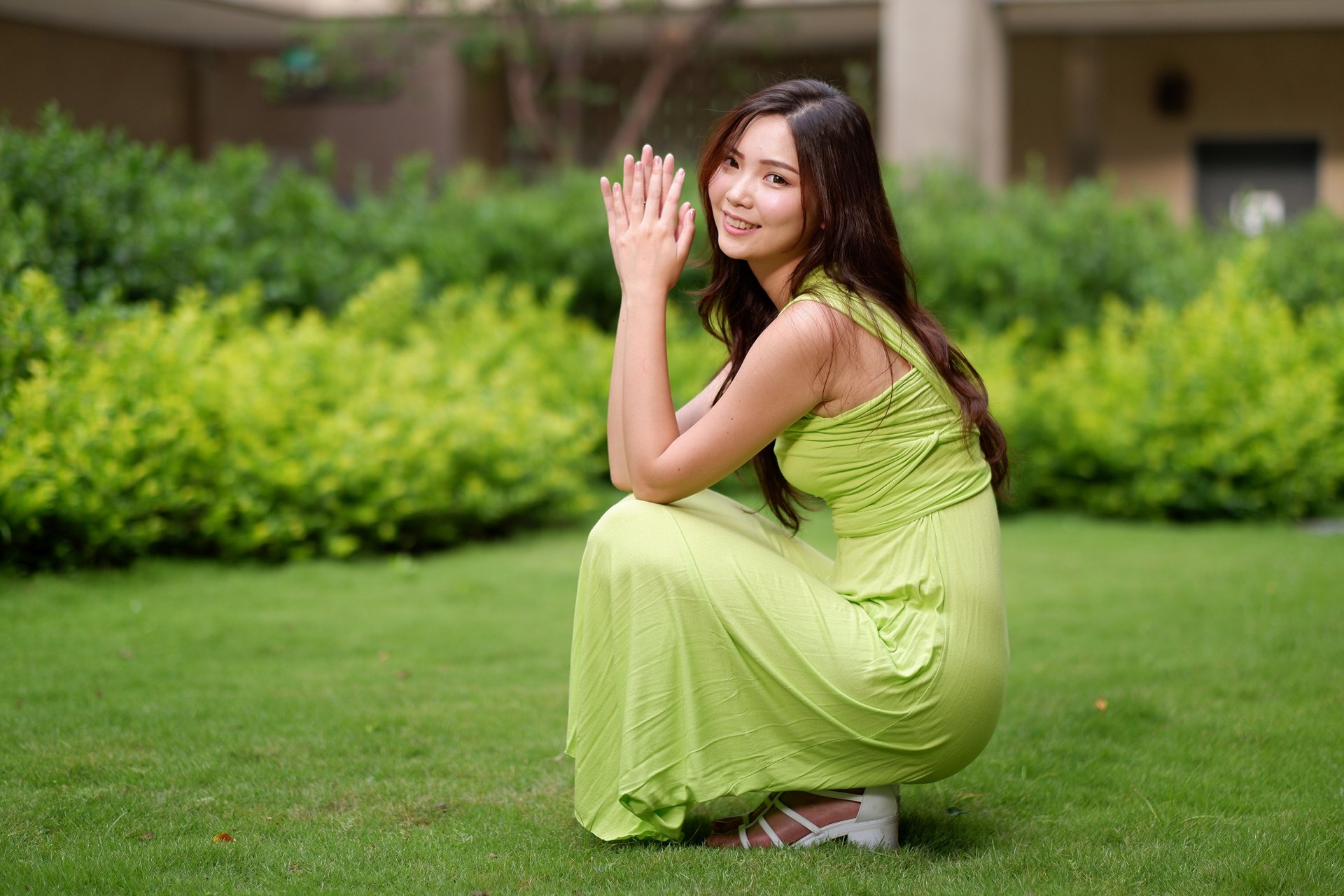 A smiling Asian woman with long brunette hair wearing a green dress squats gracefully on a lush green lawn, captured in vibrant 4K Ultra HD quality.