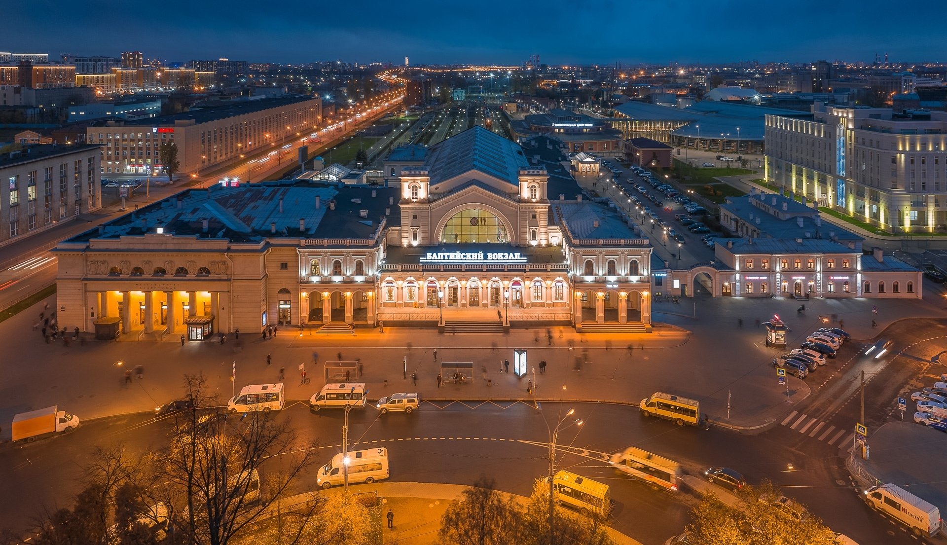 HD PC desktop wallpaper: aerial night view of Saint Petersburg's illuminated railway station, surrounding roads, buses and city buildings in a Russian urban streetscape.