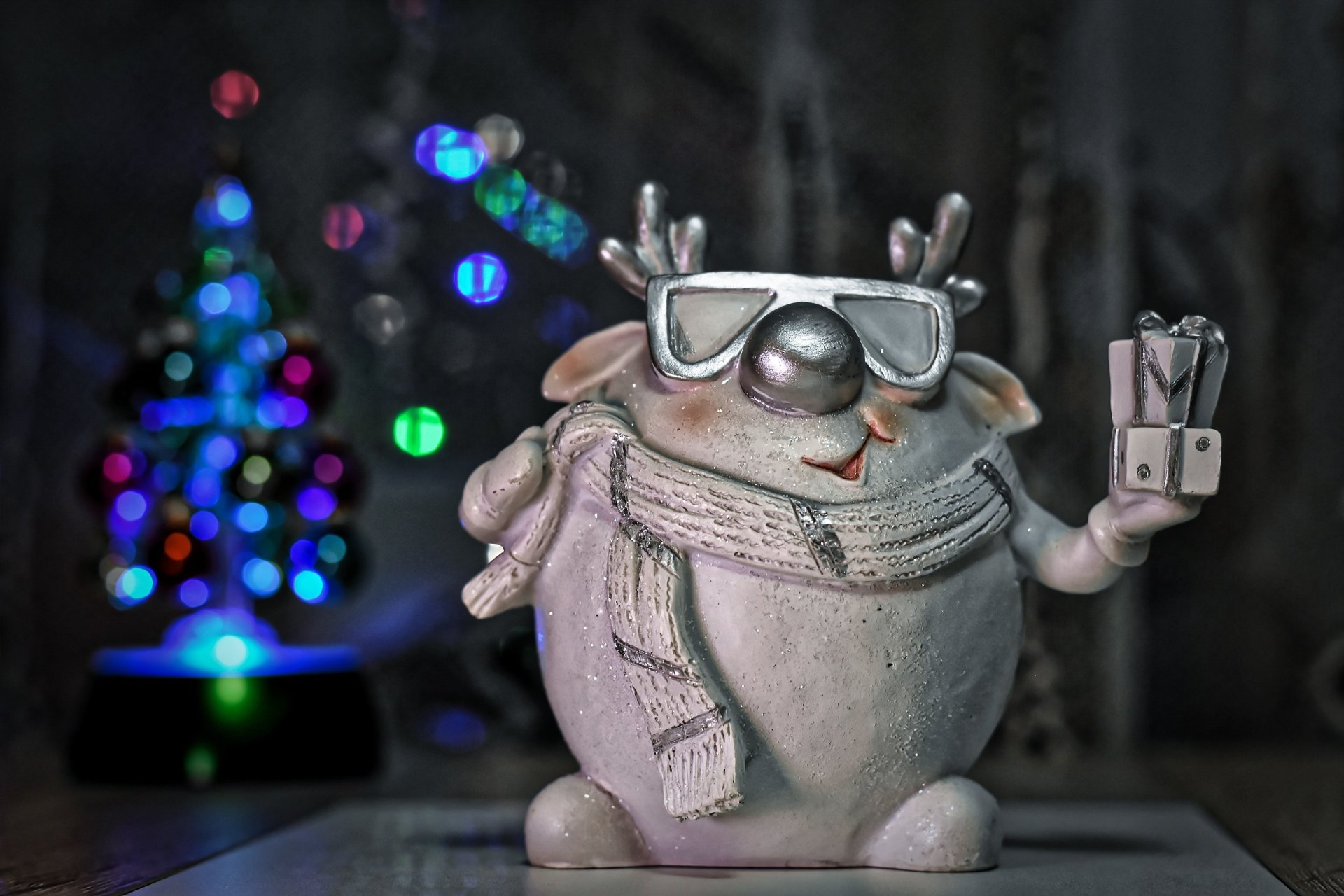 Close-up 2K Quad HD PC desktop wallpaper of a cheerful Christmas holiday figurine wearing a scarf and sunglasses, holding a small gift with a blurred decorated tree bokeh background.