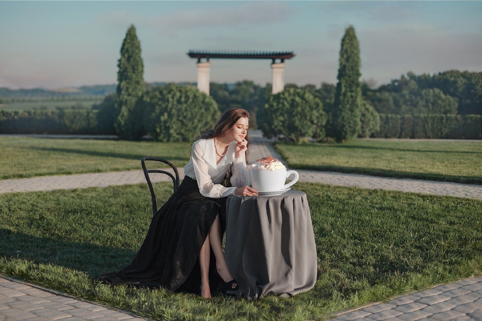 HD PC desktop wallpaper: woman model seated outdoors at a small table, legs crossed, contemplative mood as she reaches for an oversized coffee cup.