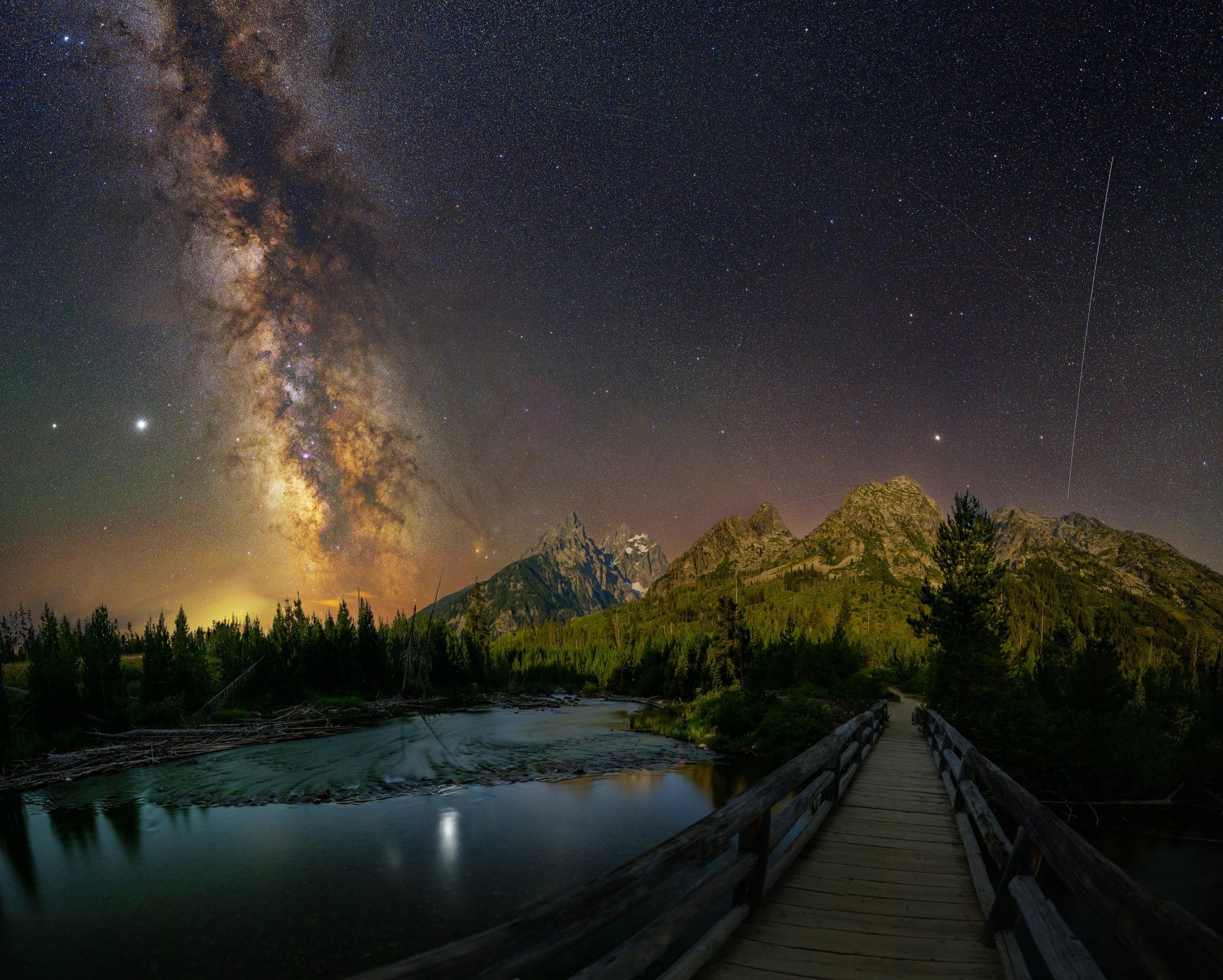 A 4K Ultra HD night scene of the Milky Way over mountains, a fir tree forest, and a lake, with a man-made bridge leading into the tranquil wilderness under starry skies.