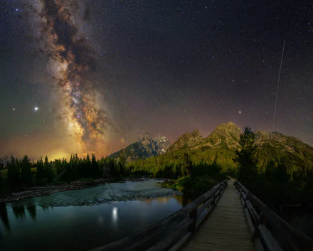 A 4K Ultra HD night scene of the Milky Way over mountains, a fir tree forest, and a lake, with a man-made bridge leading into the tranquil wilderness under starry skies.