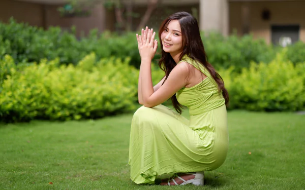 A smiling Asian woman with long brunette hair wearing a green dress squats gracefully on a lush green lawn, captured in vibrant 4K Ultra HD quality.