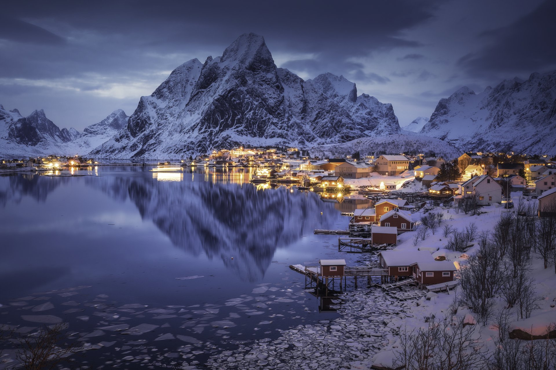 Nighttime HD desktop wallpaper of Lofoten’s snow-covered mountains reflecting in a calm lake, with village lights glowing under a cloudy sky.