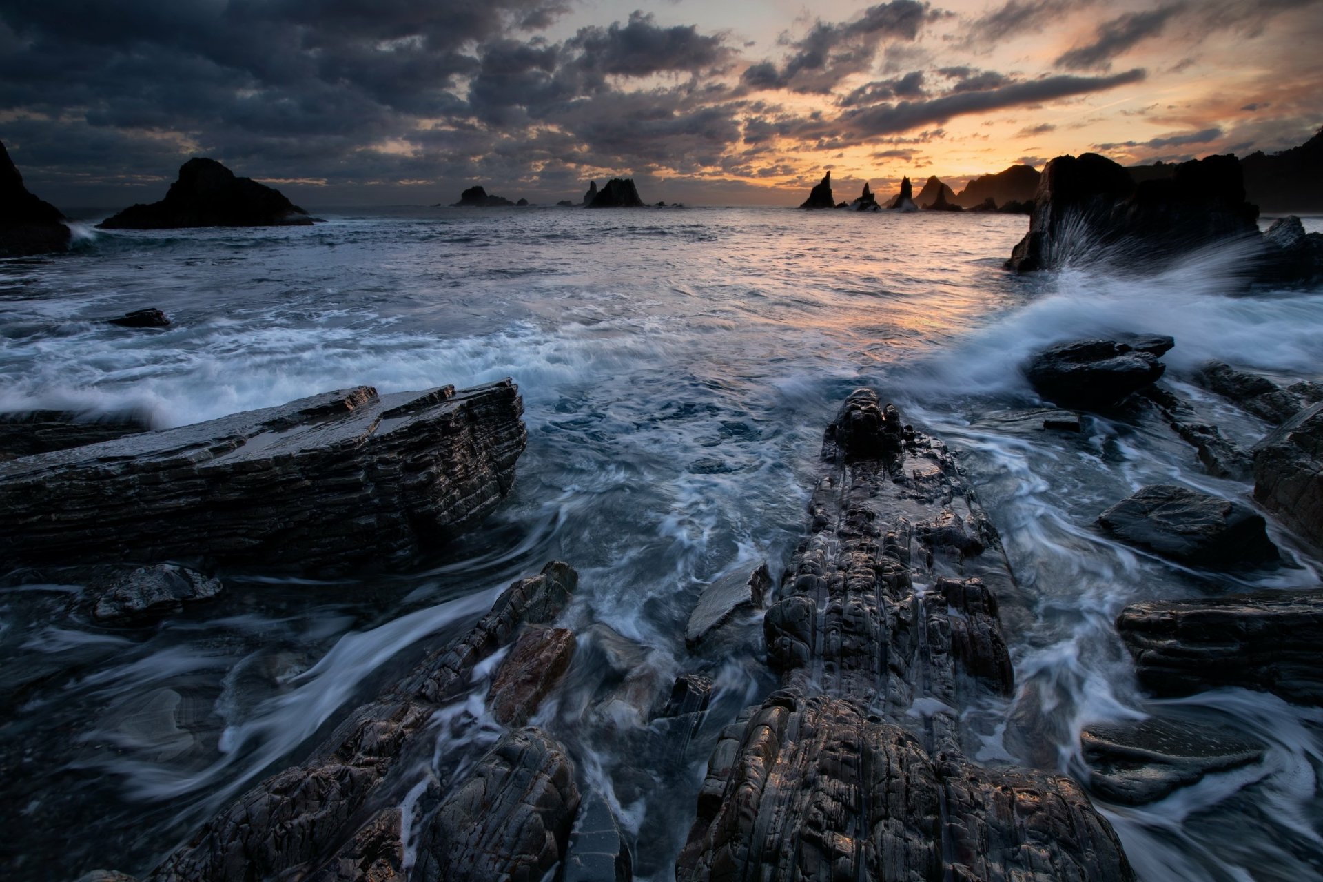 HD desktop wallpaper featuring a dramatic seascape with waves crashing against jagged rocks under a moody, cloudy sky at sunset.